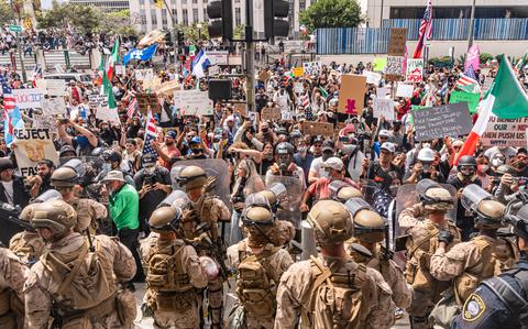 Troops in camouflage uniforms and helmets holding riot shields stand in front of a group of protesters in a plaza outside an office building.